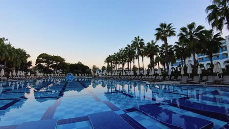 a large, tranquil resort pool lined with palm trees and sun loungers at dusk, reflecting the evening sky. ideal themes related to luxury travel, relaxation, tropical vacations, and resort lifestyle.