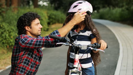 Closeup.-Portrait-of-a-pretty-girl-and-her-father-near-the-bike.-Dad-wears-a-helmet-on-the-girl's-head.-They-look-at-each-other.-Smiling.-Blurred-background