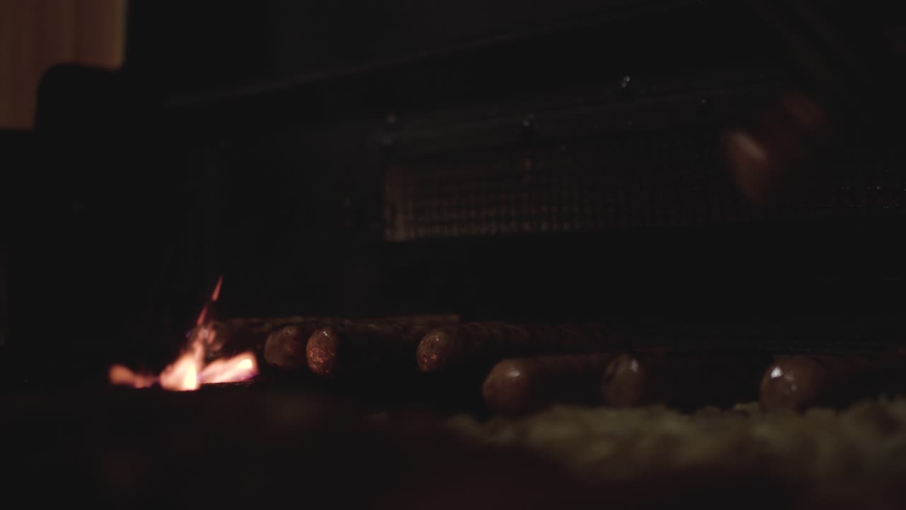 요리사는 한 쌍의 집게를 사용하여 뜨겁고 불이 붙은 그릴에서 서빙 접시로 쇠고기 소시지를 차례로 들어 올립니다.