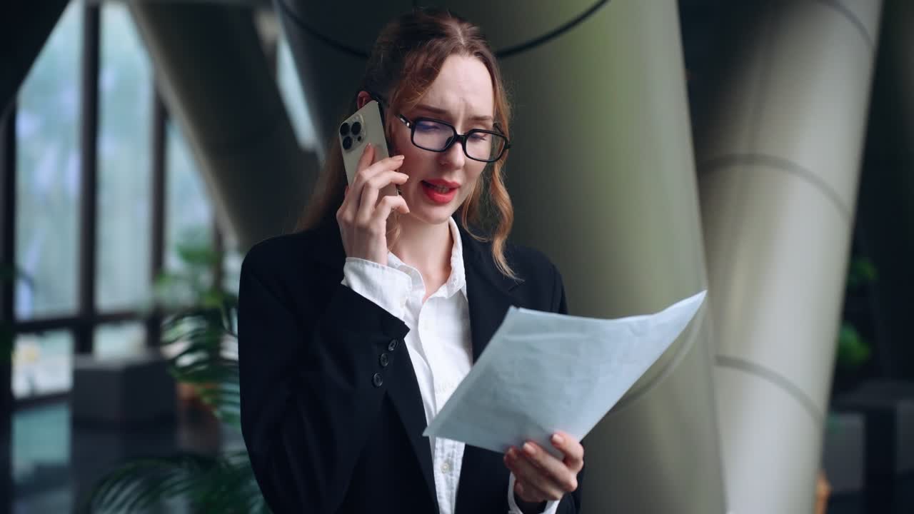 Executive woman in black suit and glasses talks on cell phone with paper work in hand standing among striking columns of corporate building