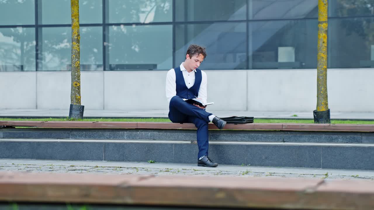 Young man in formal wear sitting on an outdoor bench, intently writing and reading on a notebook and absorbing information, representing education, focus, and scholarly pursuit, slow motion sliding