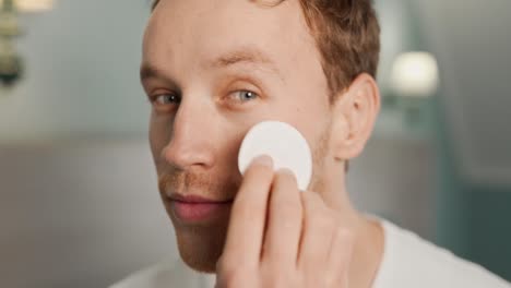 close-up portrait of a young man wiping face with cotton pad