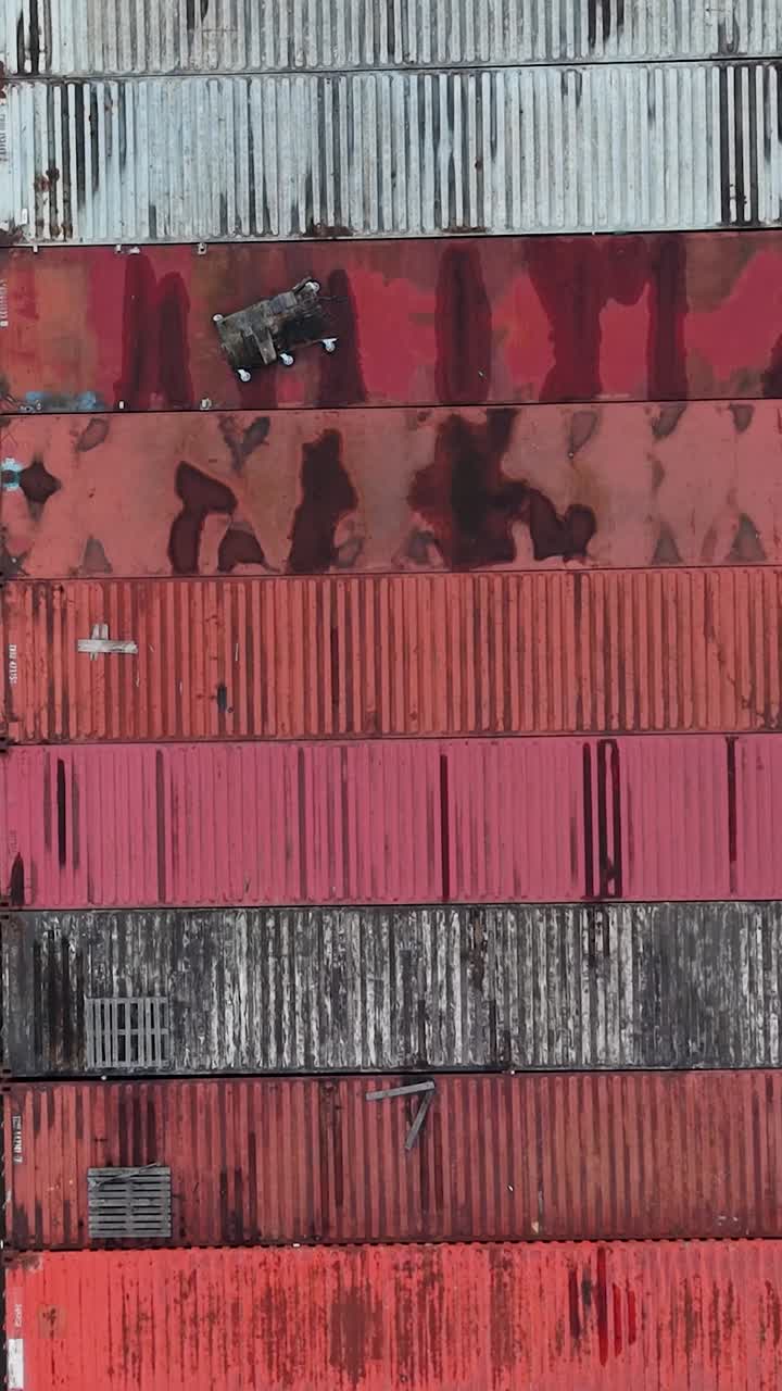 Lodret droneperspektiv ovenfra af fragtcontainere på en industrigrund, der bakker over farverige rækker ved siden af et varehustag, parkerede lastbiler, varevogne og gamle køretøjer på et travlt logistikdepot.