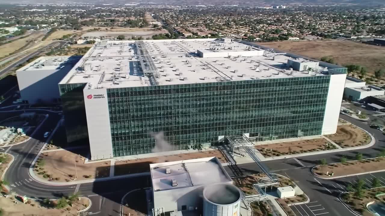 A large, modern technology facility stands prominently in an industrial area, demonstrating cutting-edge architecture. The surrounding landscape features urban elements and open spaces.