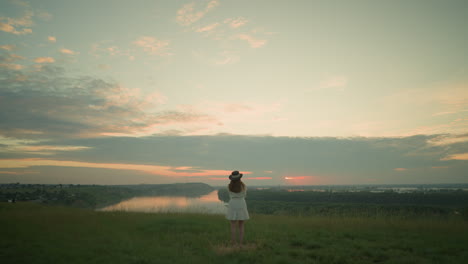 back view of a wife in a white dress and hat standing in a peaceful grassy field, gazing at a tranquil lake under a soft, pastel-colored sky during sunset, surrounded by calm and serenity