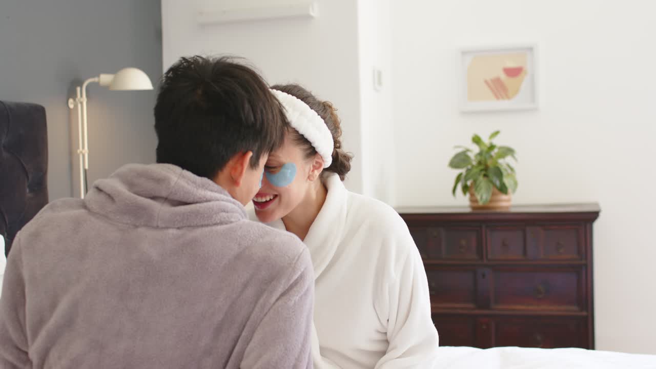 Asian man and African American woman in robes on bed talking, sharing love undereye patches visible