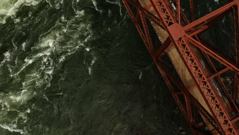 Toma-De-Un-Dron-De-Ariel-Del-Cruce-De-Un-Río-Embravecido-Bajo-Un-Puente-De-Acero-Rojo