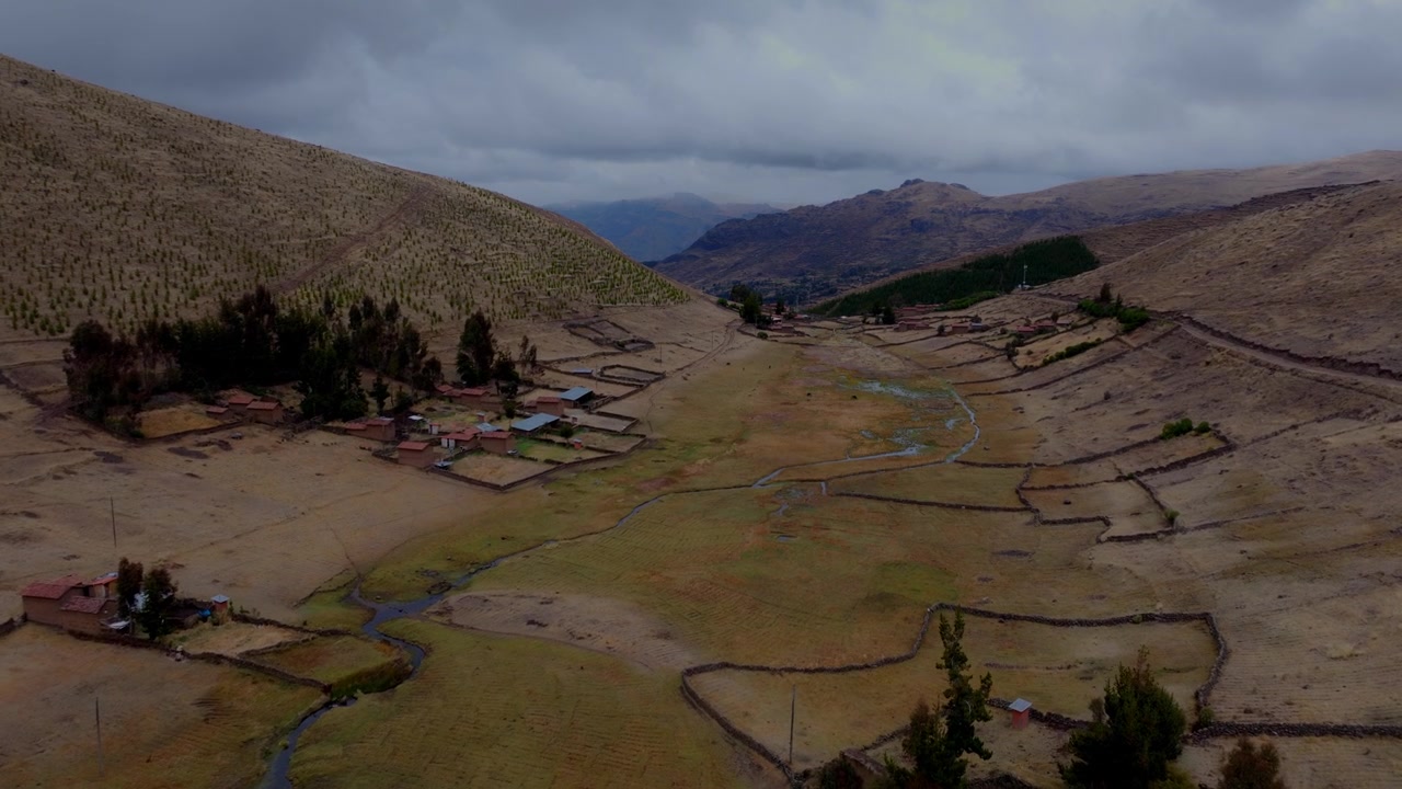 Aerial view of a remote Andean valley in Peru featuring rural stone farm plots, small rustic houses, winding streams, and rolling highlands under a cloudy sky.