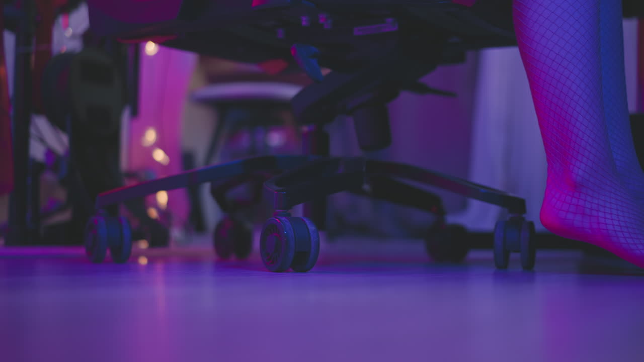 Close up leg view of lady wearing fishnet pantyhose stepping across neon lit floor toward rolling chair at gaming desk adjusts seat forward then sits down under pink purple ambient glow