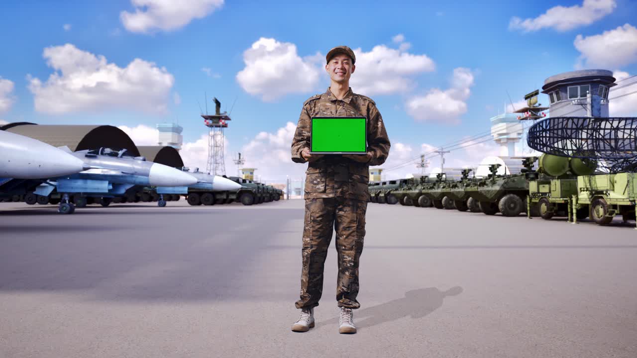 Soldato asiatico a figura intera sorridente mostra un laptop con schermo verde alla fotocamera, in piedi in un campo militare.