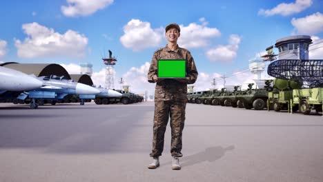 cuerpo lleno de hombre asiático soldado sonriendo y mostrando pantalla verde portátil a la cámara mientras estaba de pie en el campamento militar
