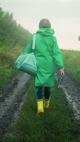 vista trasera de una mujer con un impermeable verde que lleva una bolsa de color menta y un paraguas, con botas de lluvia amarillas, caminando por un camino fangoso rodeado de vegetación alta, árboles y un cielo brumoso