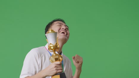 close up of asian man with a gold medal looking at a gold trophy then celebrating winning as the first winner on green screen background in the studio