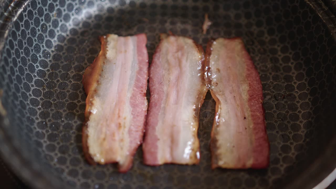 Three strips of bacon cooking and sizzling in a frying pan. Static close up view.