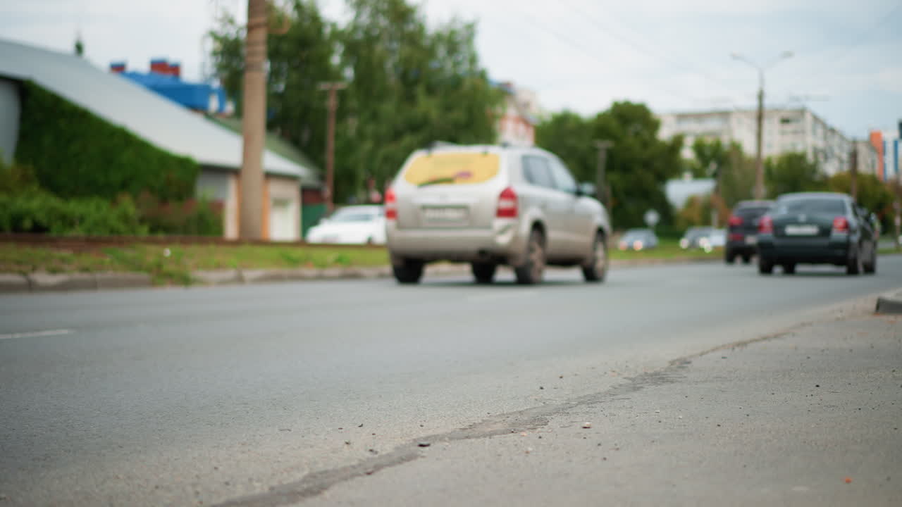 Cars Passing Along Cracked Asphalt Road With Vans And Sedans, Daytime Suburban Intersection Framed By Trees And Buildings, Dynamic Traffic Flow With Brake And Turn Actions, Visual Emphasis On Roadside