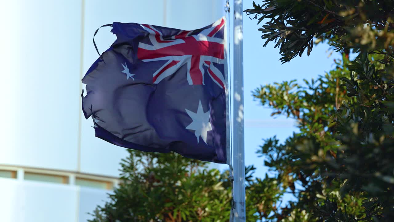 A tattered Australian flag waves vigorously against a clear sky, surrounded by lush greenery in Bellarine, Victoria