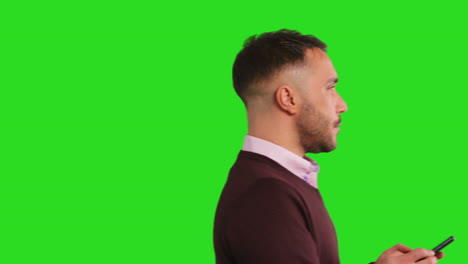 Close-Up-Studio-Shot-Of-Walking-Businessman-Using-Mobile-Phone-To-Message-Or-Browse-Online-Standing-Against-Green-Screen