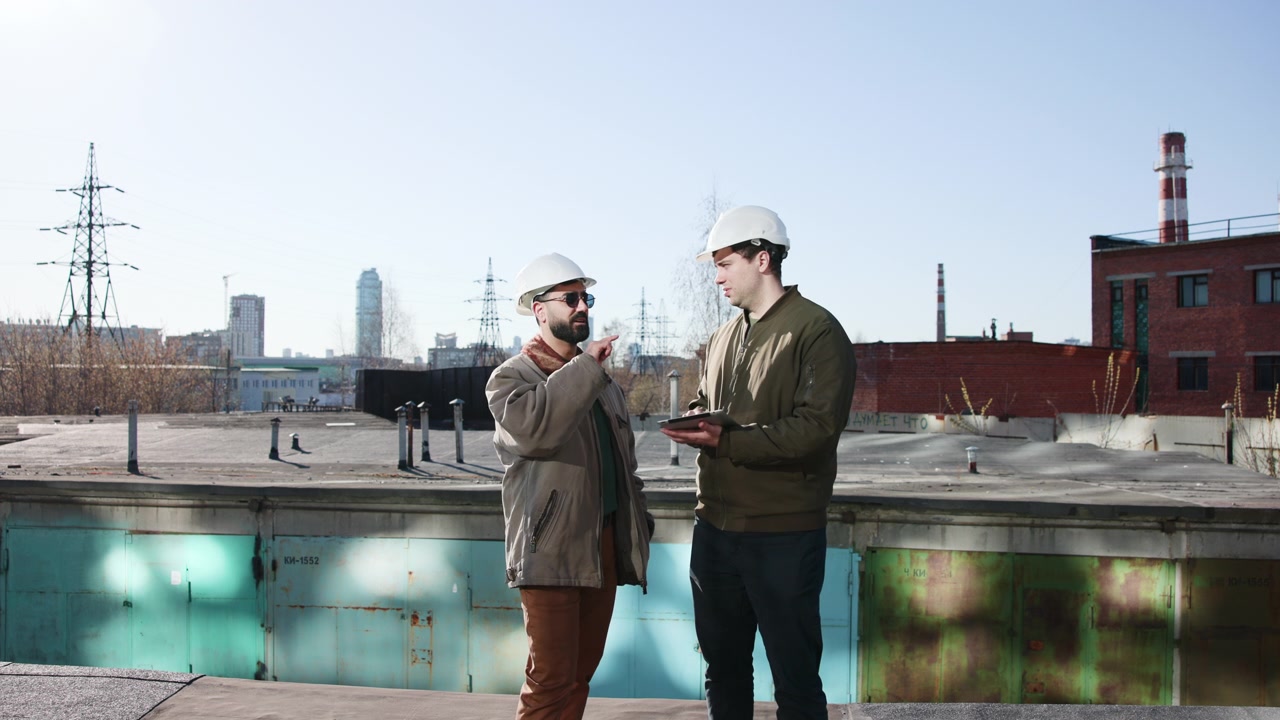 Two Engineers On Rooftop Reviewing Tablet, Both Wearing White Helmets And Jackets, Urban Skyline And Power. Media