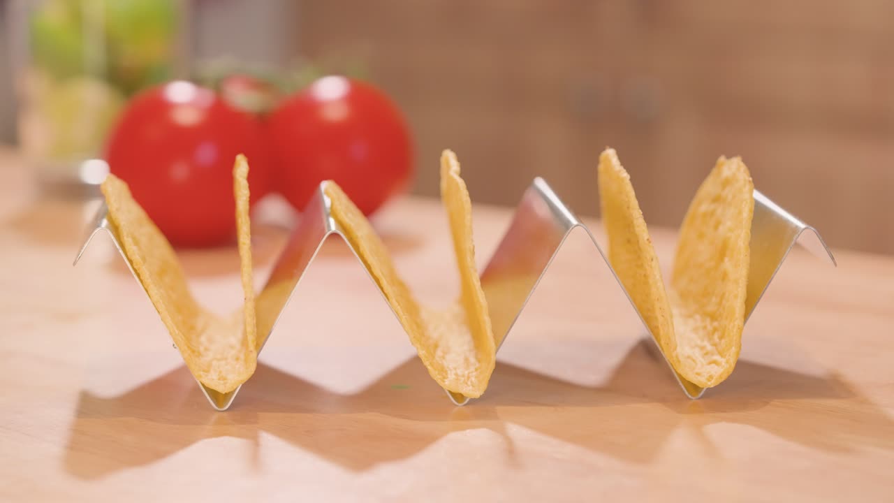 Close-up of an empty stainless steel taco stand as a hand adds 3 corn taco shells one by one, with tomatoes and limes in the background.