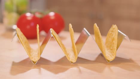 close-up of an empty stainless steel taco stand as a hand adds 3 corn taco shells one by one, with tomatoes and limes in the background.
