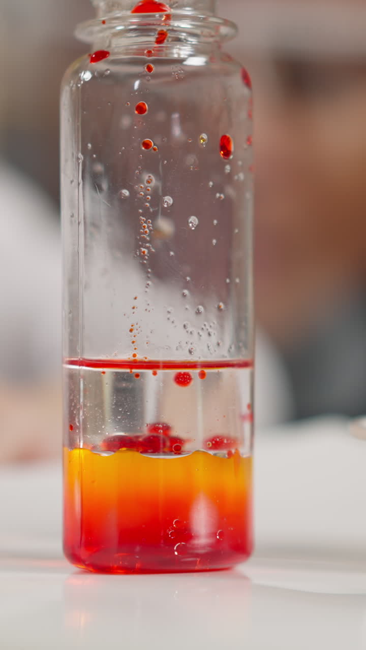 Little schoolgirl drips bright red oil paint onto transparent liquid surface performing chemical experiment with black teacher in lab extreme closeup