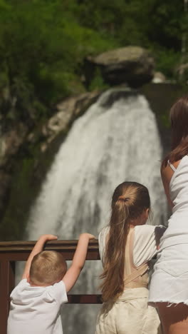 little children spellbound by waterfall and mother turns back on park bridge. woman with cute kids travel to look at river cascade at wilderness resort