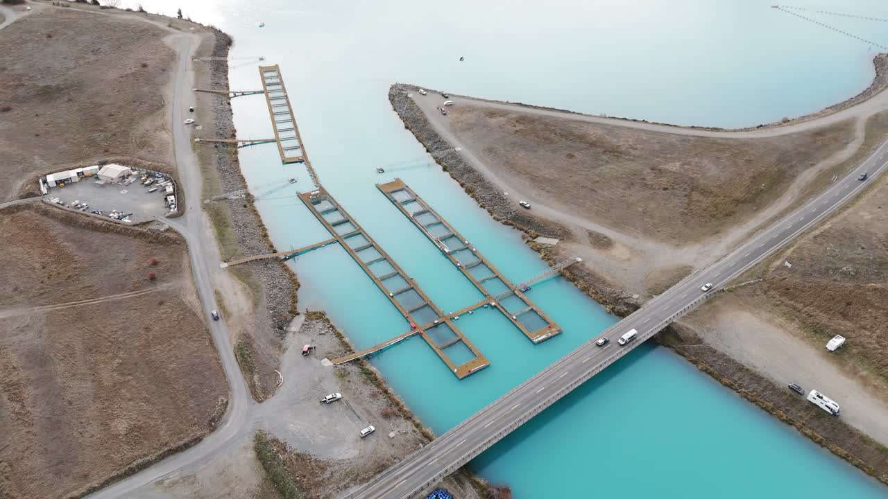 Slow descending aerial approach over High Country Salmon farm in Twizel, with pens floating in turquoise water and cars visible crossing the adjacent bridge