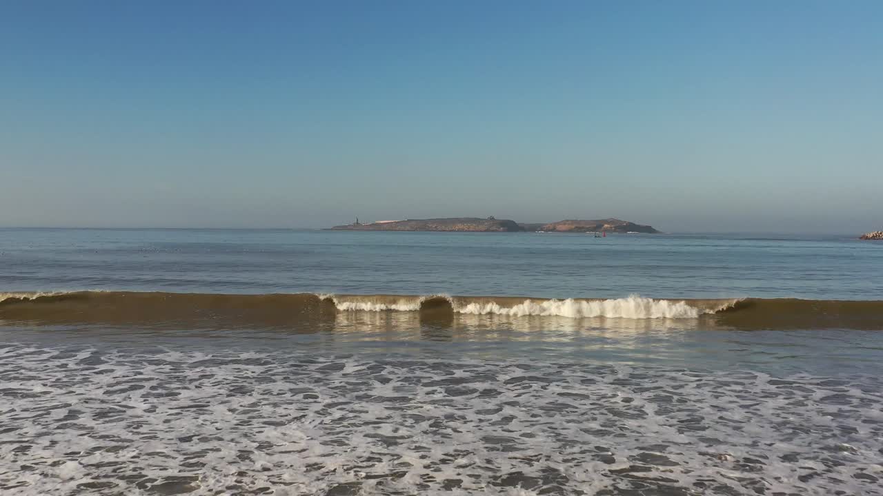 Peaceful view of gentle ocean waves rolling onto a golden sandy shore with a rocky island visible in the background under a clear blue sky