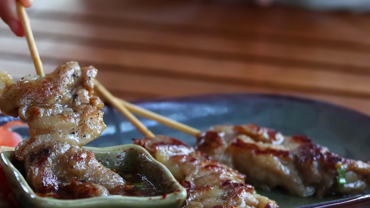 Close-up of grilled skewers being dipped into a flavorful sauce on a dark plate.