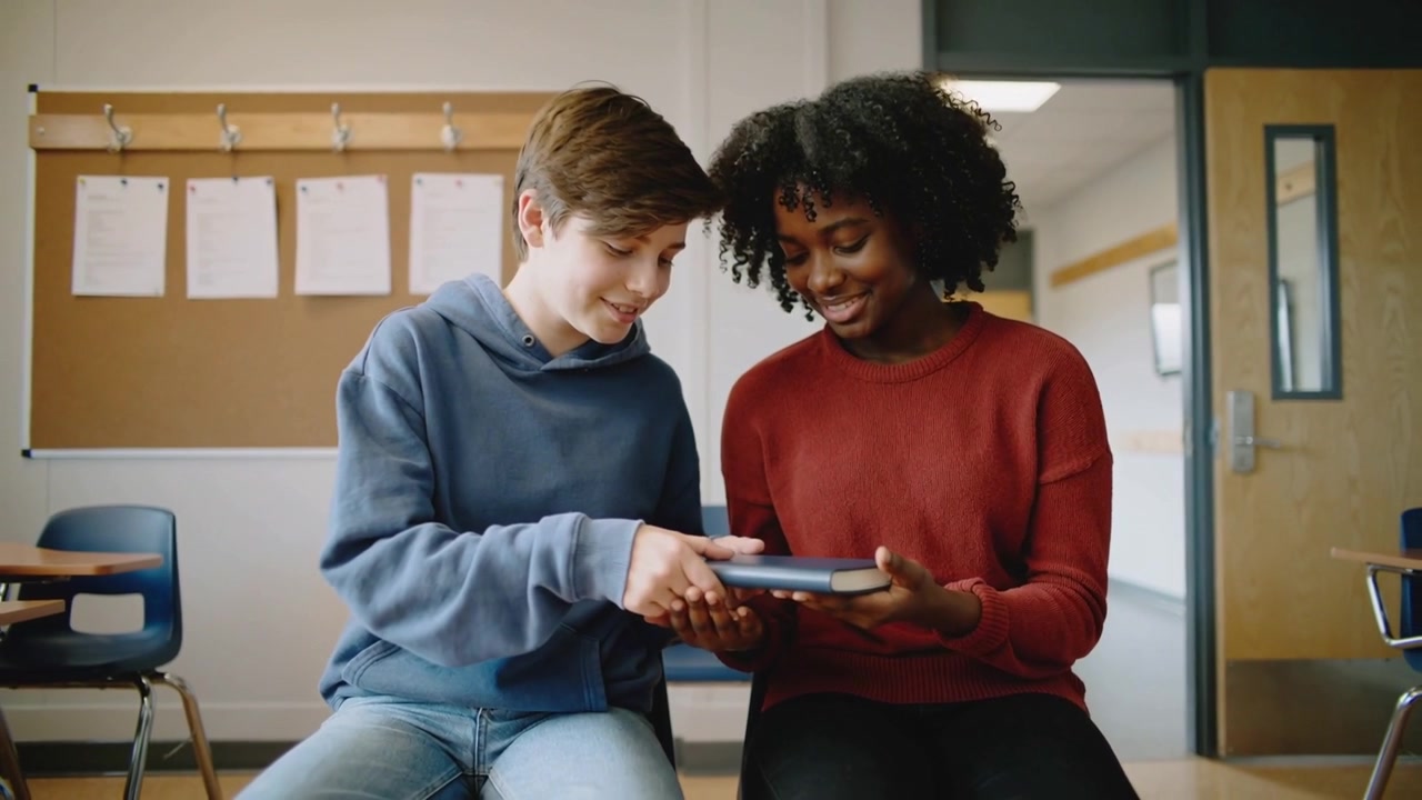 Reaching male offering blue book to female in classroom, wearing blue hoodie, red sweater, reading