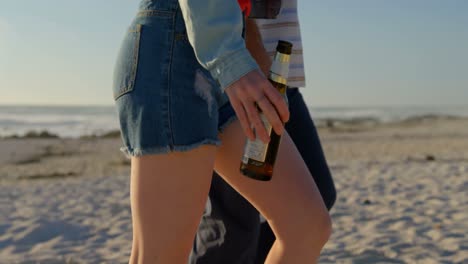 Side-view-of-couple-walking-with-beer-bottle-at-beach-on-a-sunny-day-4k