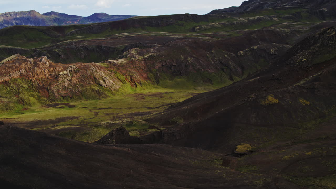 Pull back drone shot of a mountain valley in Iceland, as bright green moss and volcanic black sand contrast nicely in the sun illuminated valley.