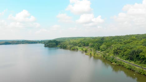 Drohnenflug-In-Den-Horizont-über-Einem-See-An-Einem-Blauen,-Sonnigen,-Weißen,-Bewölkten-Himmel,-Während-Ein-Auto-Die-Landstraße-4k-Hinunterfährt