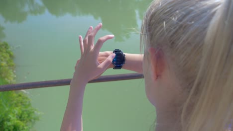 a-young-blonde-woman-clicks-and-scrolls-on-her-connected-watch-on-a-bridge-over-a-river-and-a-park