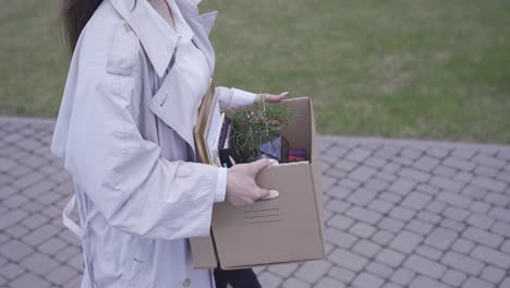 una mujer joven camina frente a un edificio de oficinas. ella lleva sus cosas en una caja. ella ha dejado su trabajo. ella esta sonriendo.