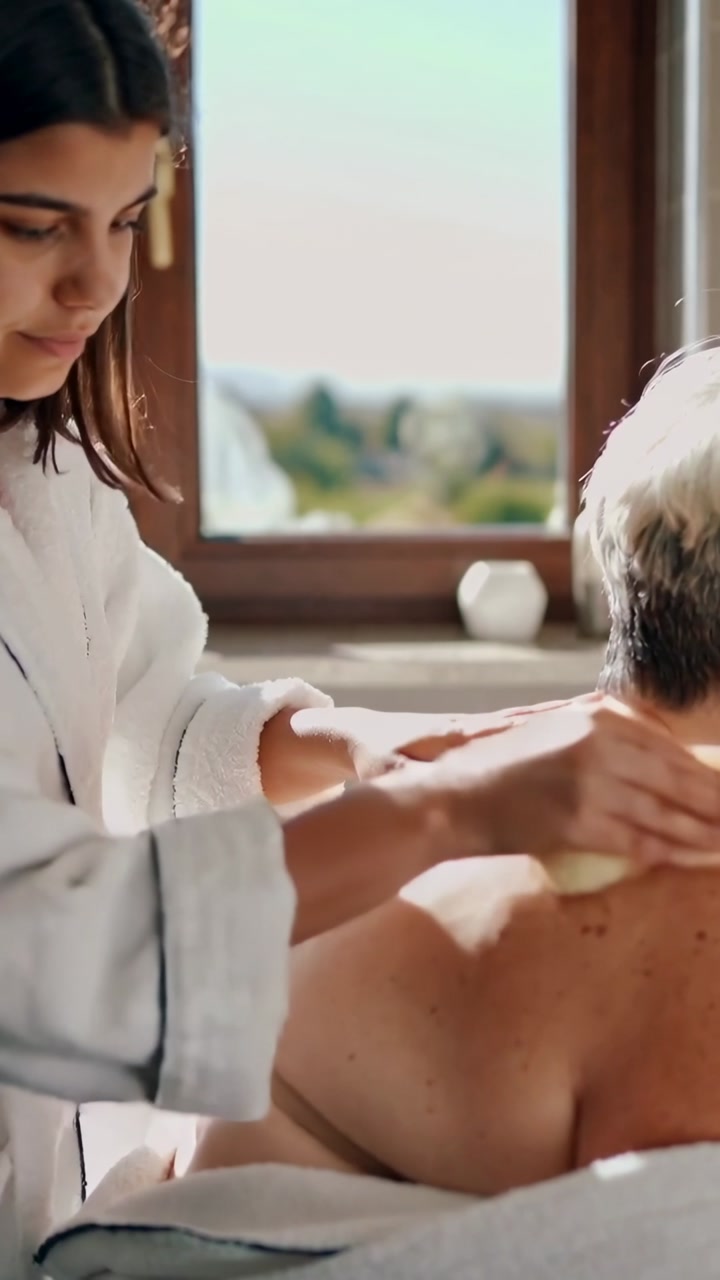 Vertical video: Placing sponge, caregiver white robe rubbing senior back on towel at home for care