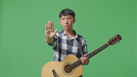 asian teenager with guitar making stop gesture while standing on green screen background