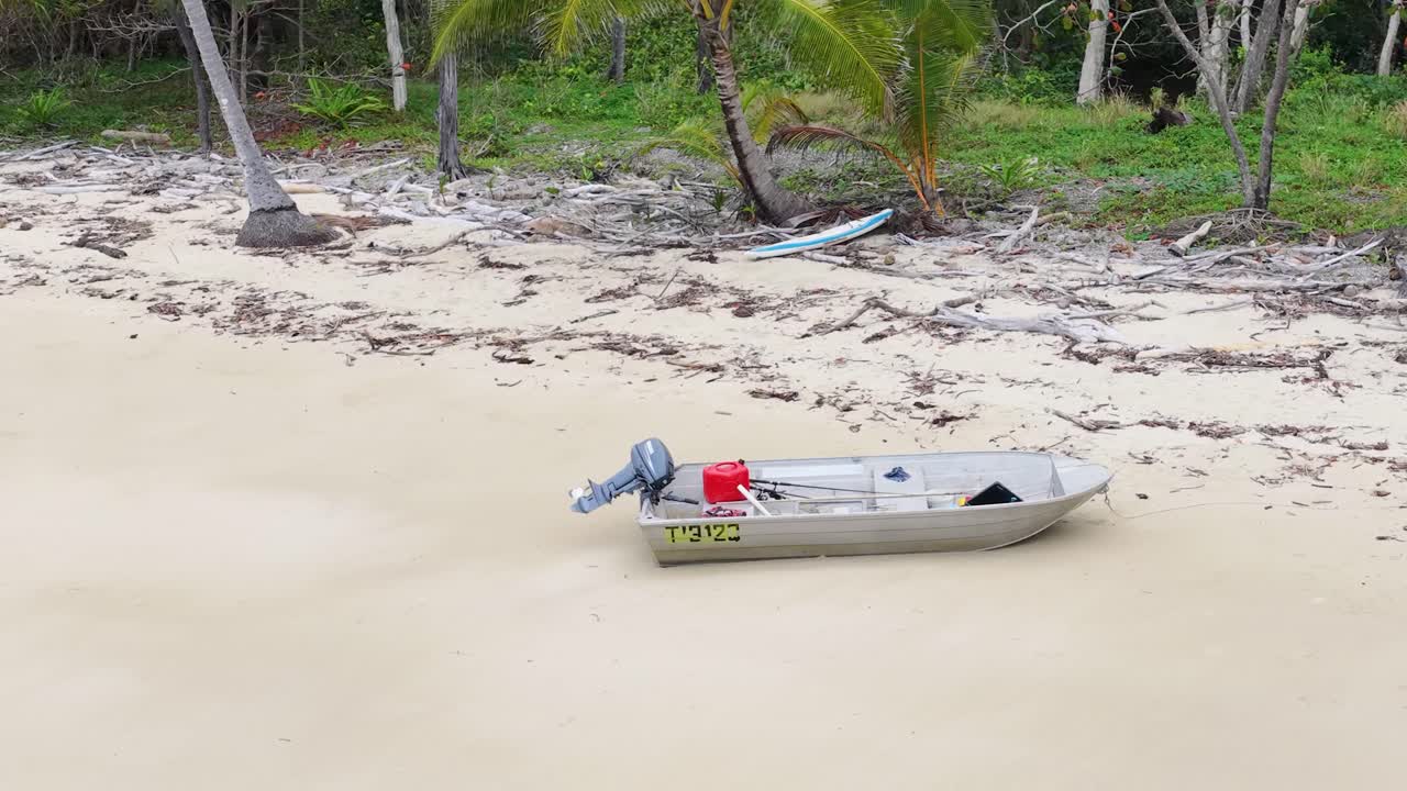 Aluminum boat with outboard motor on sandy beach, rainforest backdrop, overcast daylight, aerial view