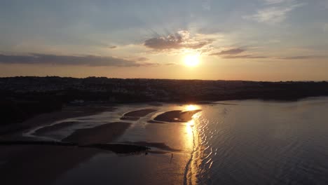 Sunset-rays-beam-fiery-golden-colours-across-relaxing-beach-ocean-coastline-aerial-view-slow-rising-pull-back