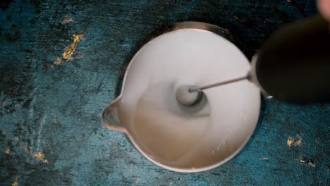 top-view-coffee-creamer-with-electric-milk-frother-stick-mixing-milk-until-it-bubbles-up-on-blue-background