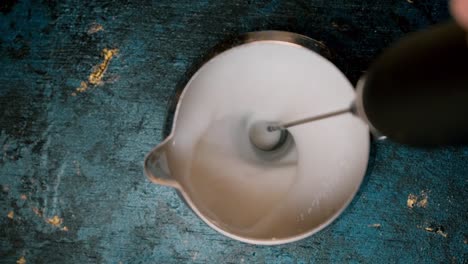 top view coffee creamer with electric milk frother stick mixing milk until it bubbles up on blue background