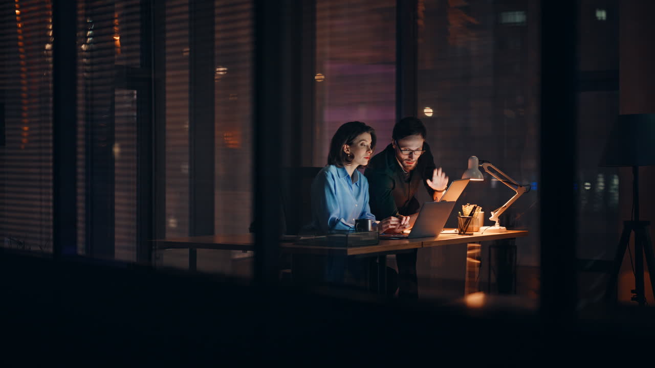 Two professionals cooperating project overtime working at laptop in lamp light