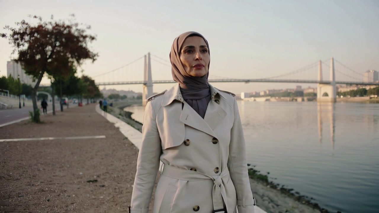 Kvinde i beige trenchcoat og hijab går mod kameraet ved en flod, bro synlig