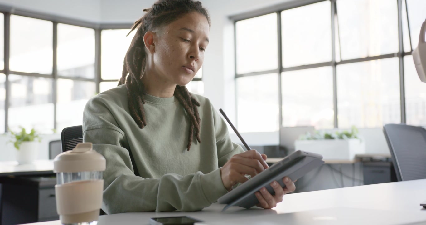 Nonbinary designer starting sketch sitting at desk by window, holding stylus refining art on tablet