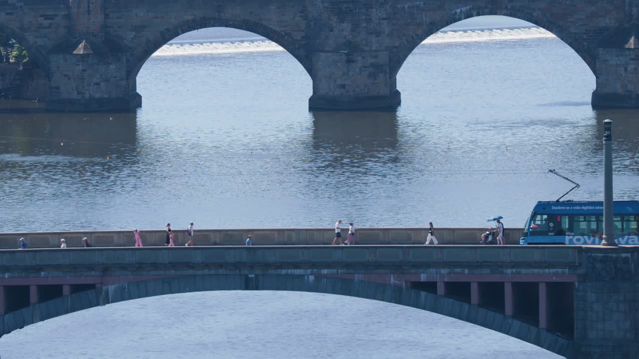 Blue tram travels across bridge with pedestrians, river reflections, and historic arches in daylight