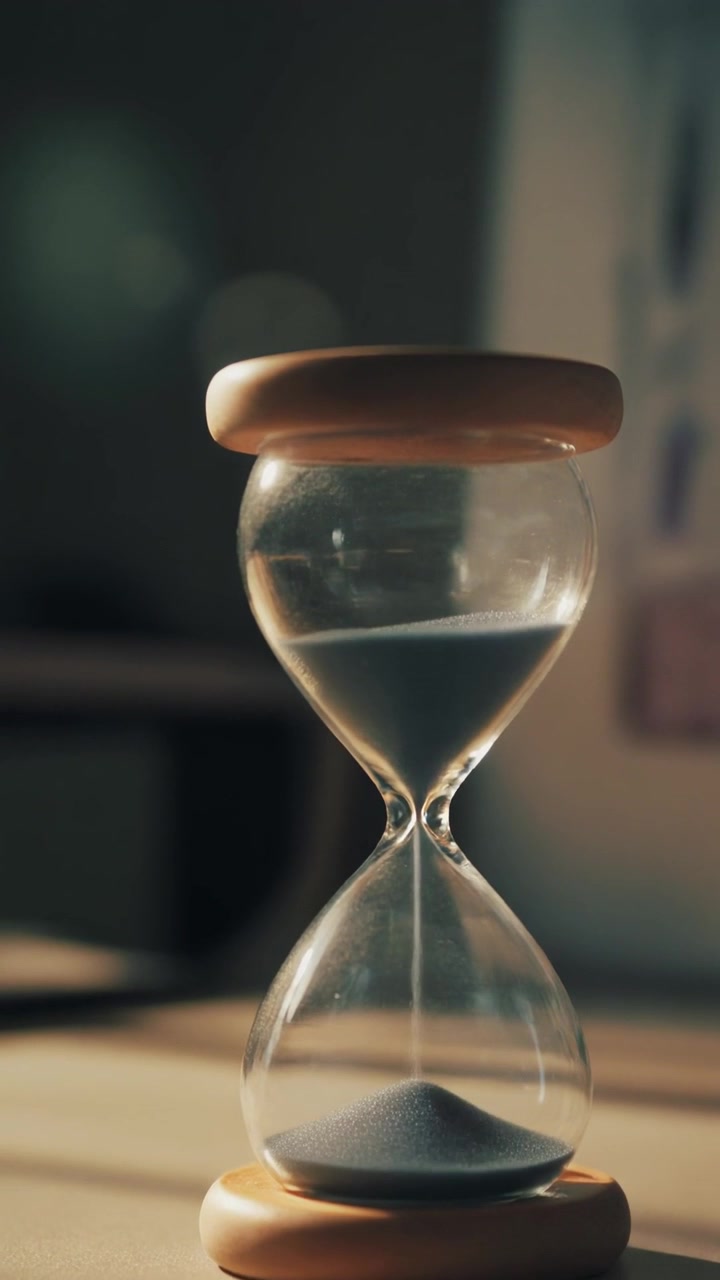 Hourglass with wooden caps filled with dark grey sand on wooden table in soft sunlight