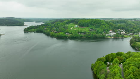 An-aerial-panorama-of-a-lakeside-village-with-houses-nestled-among-green-hills-and-trees,-reflecting-in-the-water