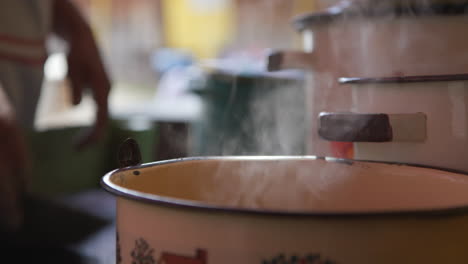 Un-Primer-Plano-De-Una-Olla-Hirviendo-En-Una-Estufa-Con-Una-Mujer-Cocinando-Tortillas-En-El-Fondo