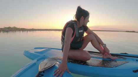 young woman sitting on sup paddle board over still lake water during sunset smiling at camera