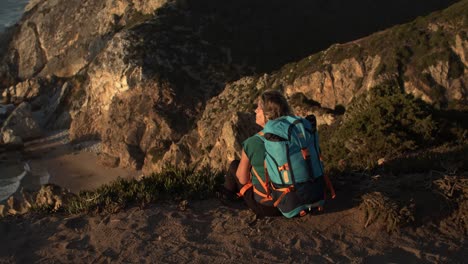 Vista-Posterior-De-Una-Mujer-Con-Mochila-Sentada-En-El-Acantilado.